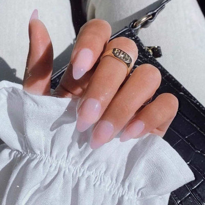 A hand with long, almond-shaped nails in light pink—Cloud 9 by glamnetic—rests on a textured black bag. A gold ring and ruffled white sleeve complete the look, highlighting a chic and elegant manicure.